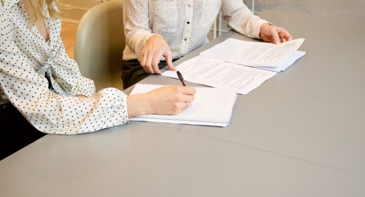 woman signing on white printer paper beside woman about to touch the documents